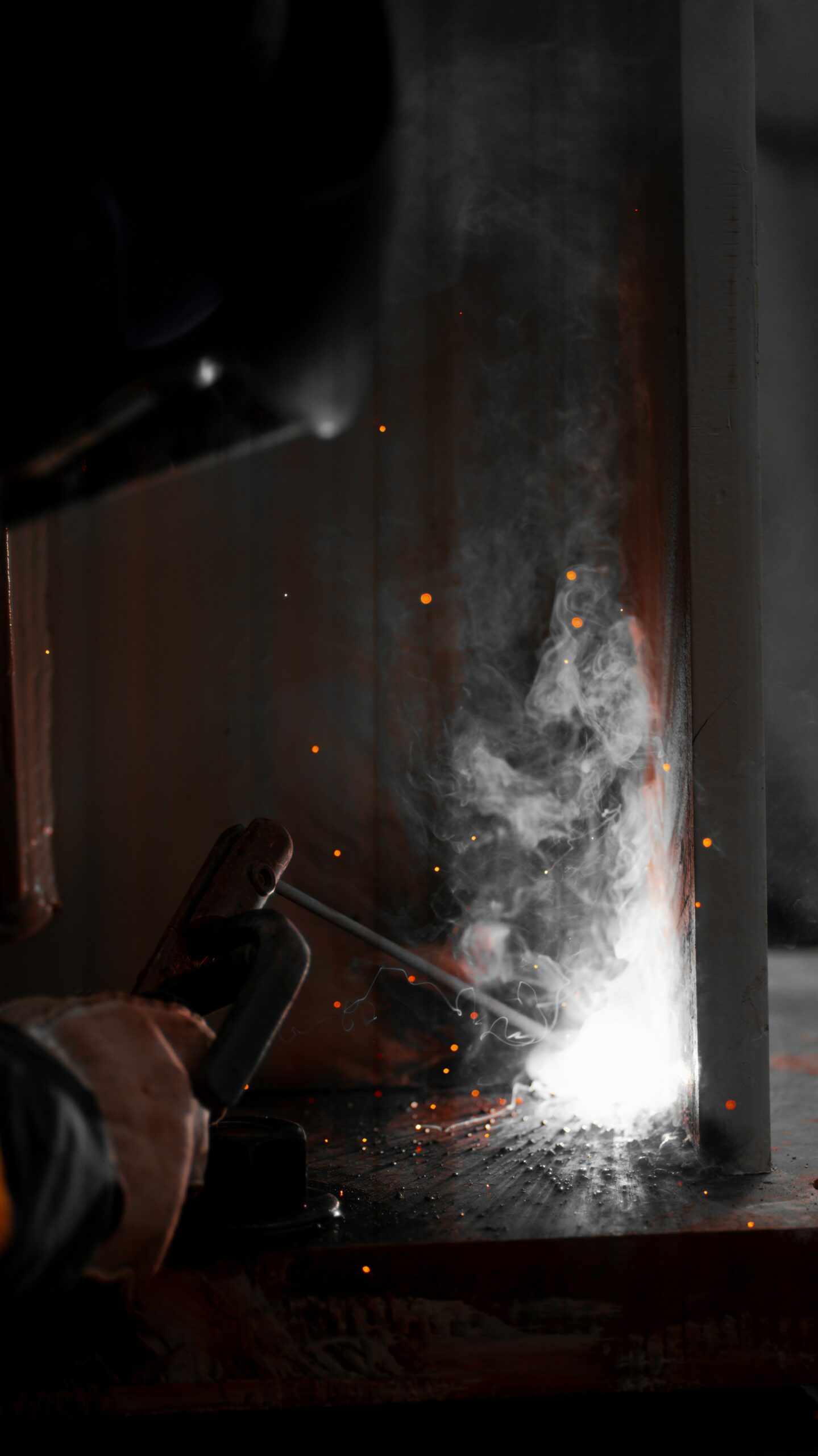 Close-up of welding in progress, with glowing sparks and rising smoke as a welder uses a handheld electrode on a metal surface.