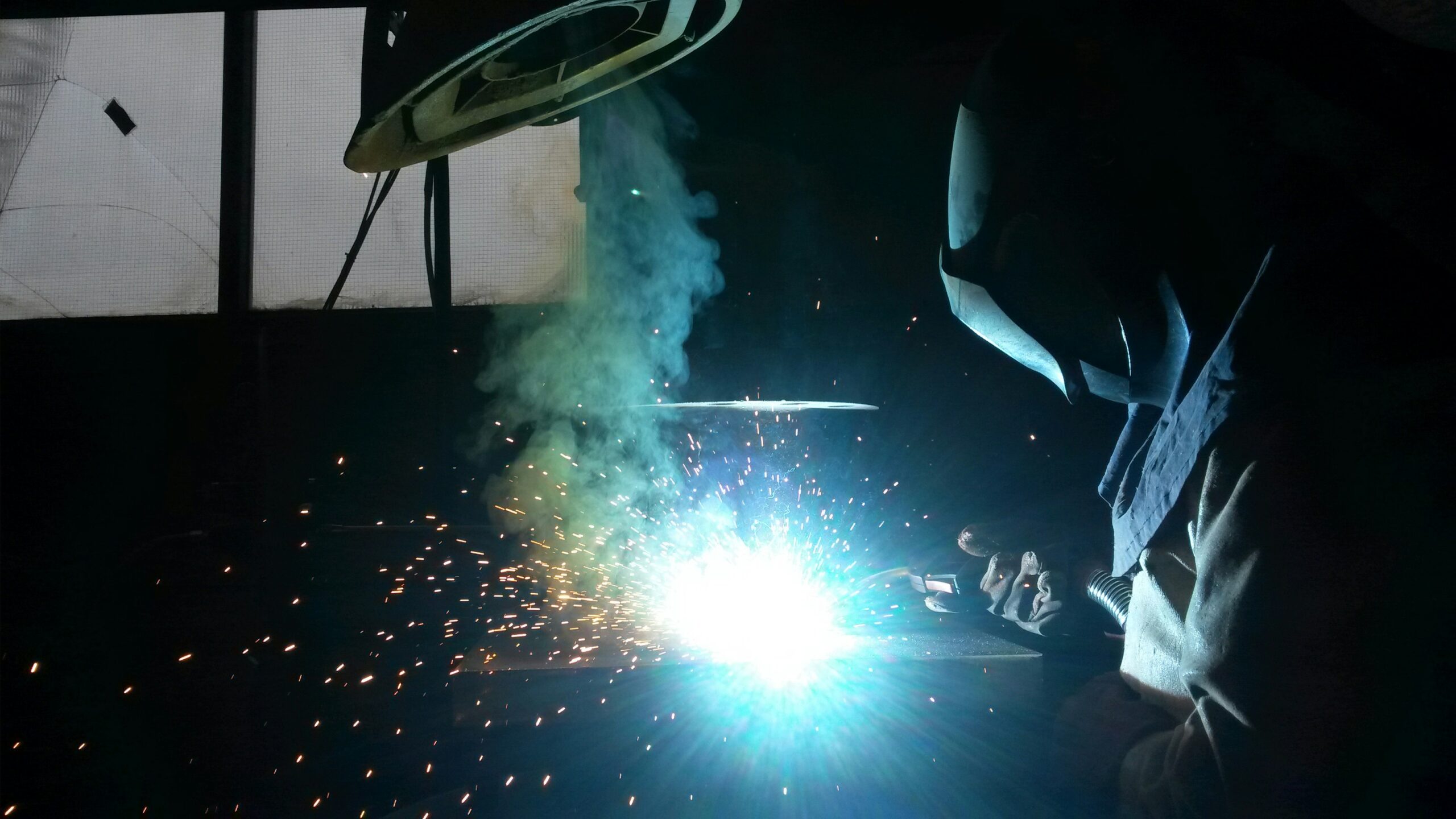 Person welding metal with protective helmet and gloves, bright sparks and blue light illuminating the workshop.